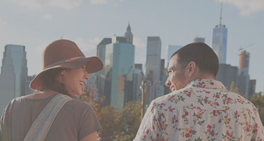 Man in floral shirt and woman in hat talking smile at each other in front of a city skyline.