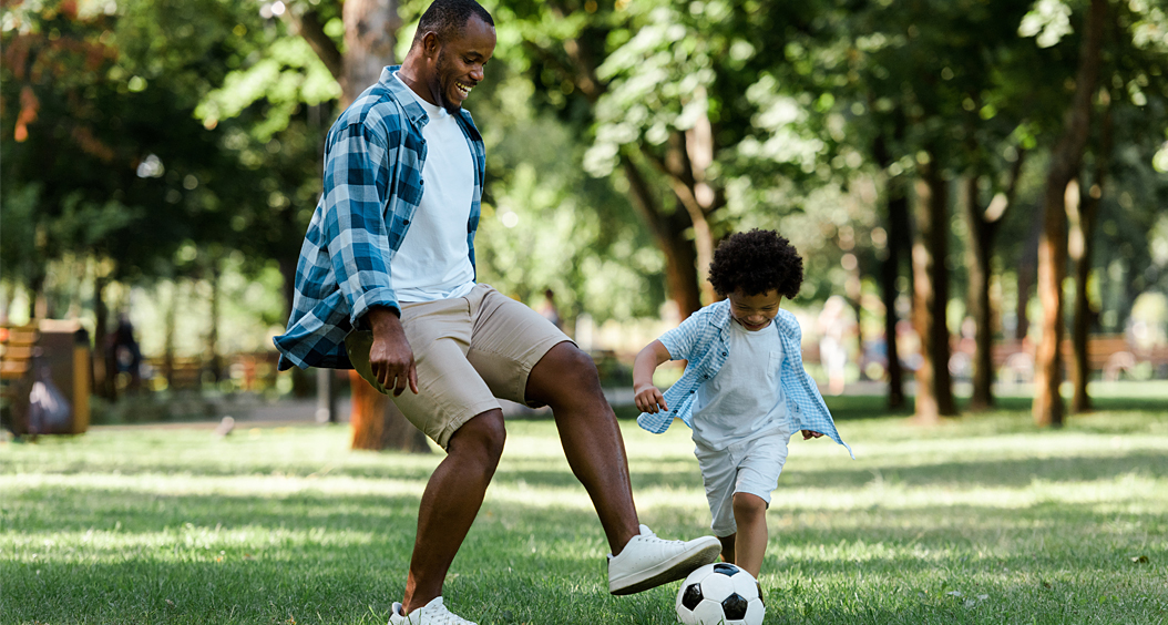 Happy African American man playing soccer with son outside.