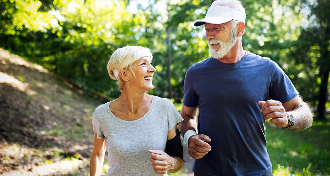 Senior couple smile at each other while jogging in a wooded area.