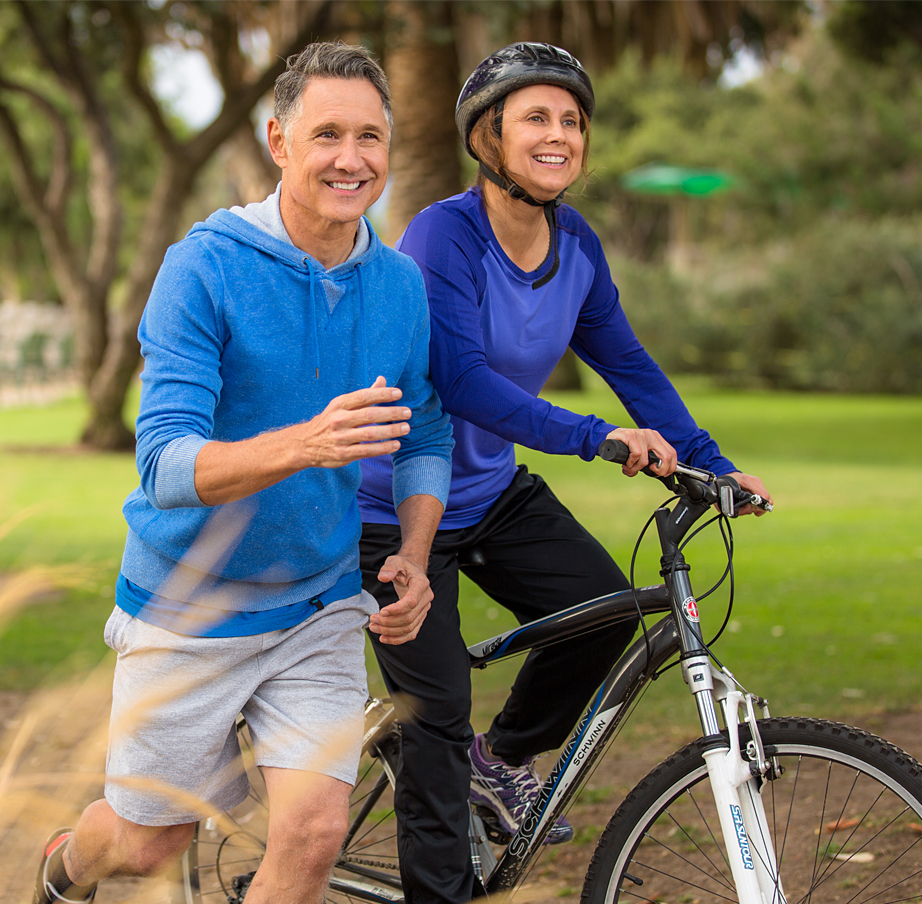 Senior female rides a bicycle next to her jogging husband.