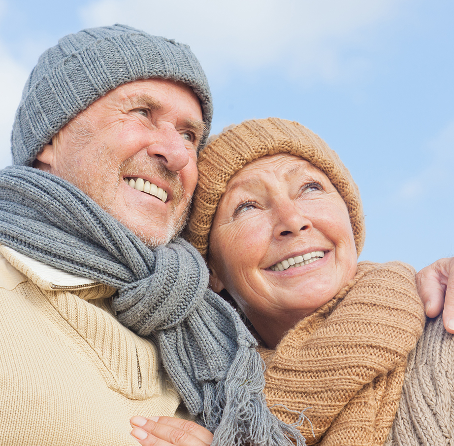 Senior couple in sweaters, knit caps, and scarves smile and embrace against a blue sky.