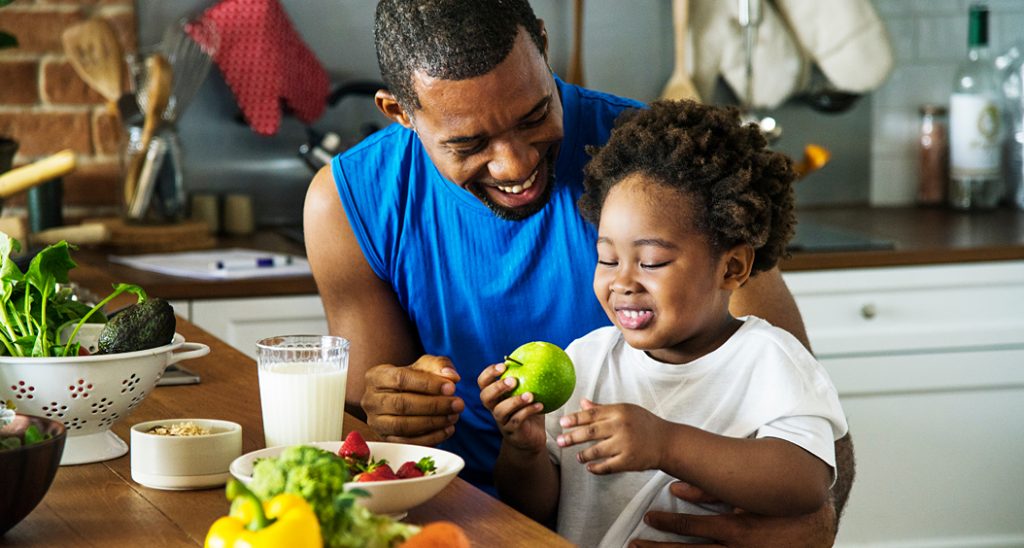African American girl eats fruit while sitting on her smiling father's lap.