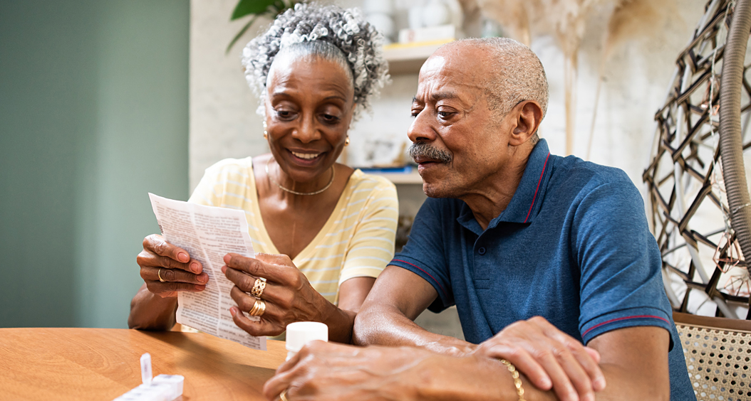 Senior African American couple look at prescription drug information sitting at a table inside.