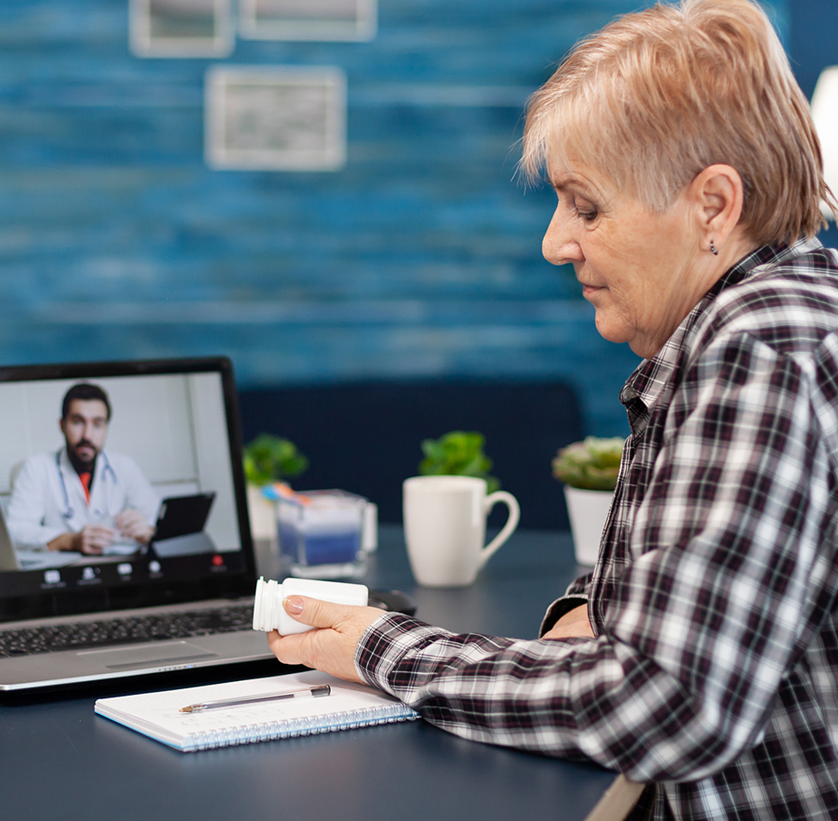 Senior woman examines prescription bottle while talking with a doctor via laptop.