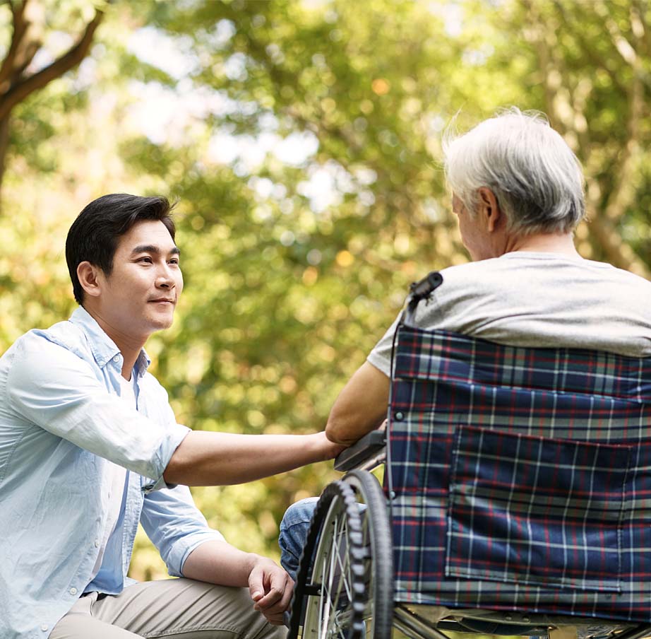 Asian son talking to and comforting wheelchair bound father outside.